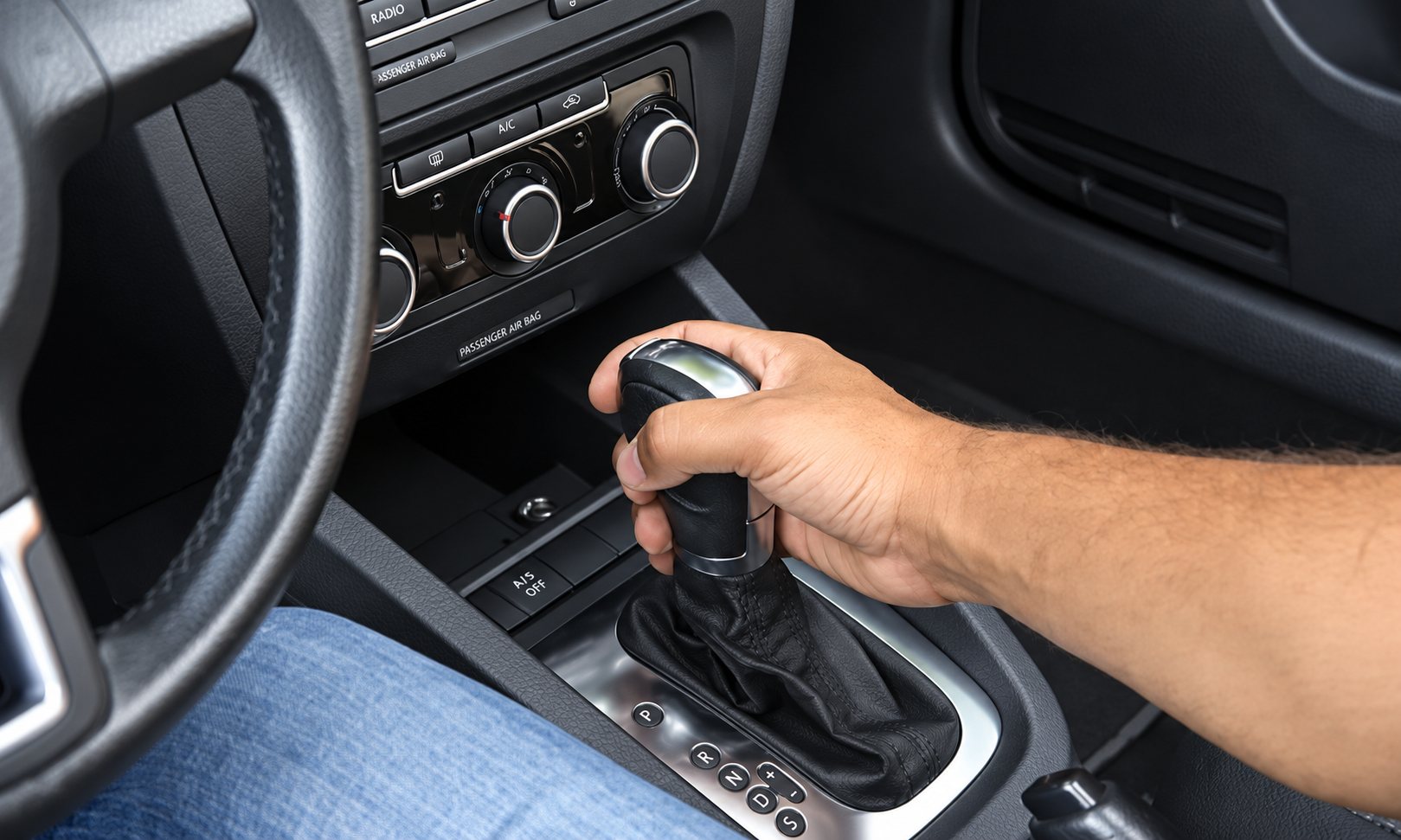 Une main d’homme tient le levier de vitesses d’une boîte automatique dans l’habitacle d’une voiture, près de la console centrale et des commandes de climatisation.